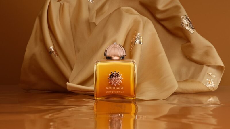 Amouage Overture41 bottle on a reflective amber surface in front of draped golden fabric