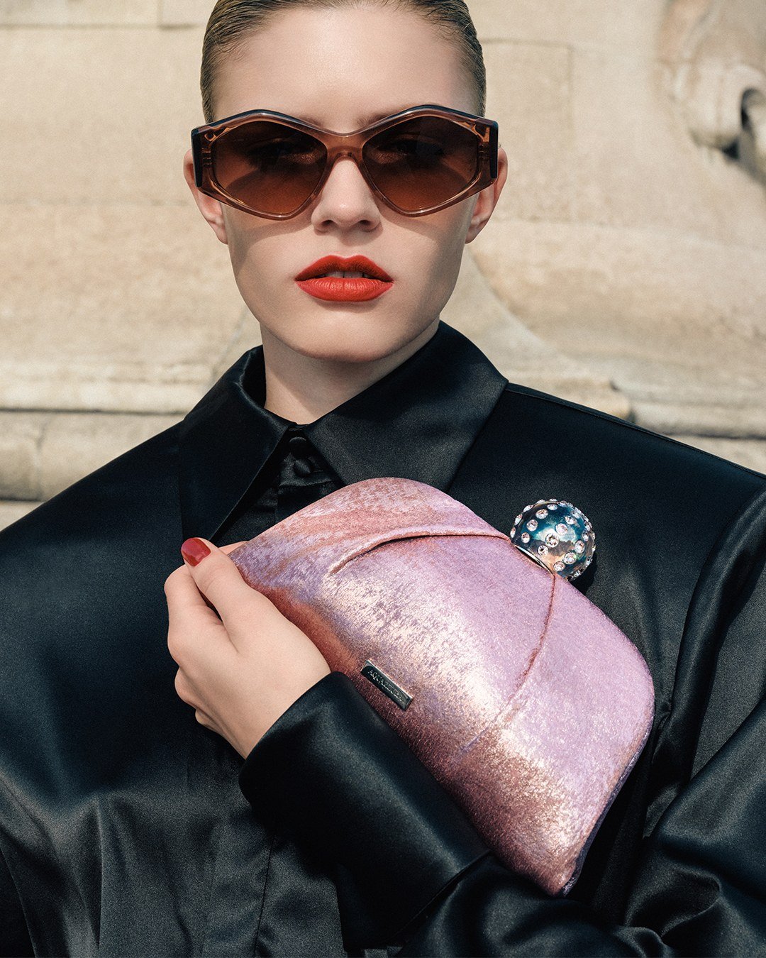 Model in sunglasses holding a shimmering pink Aquazzura clutch against a black shirt