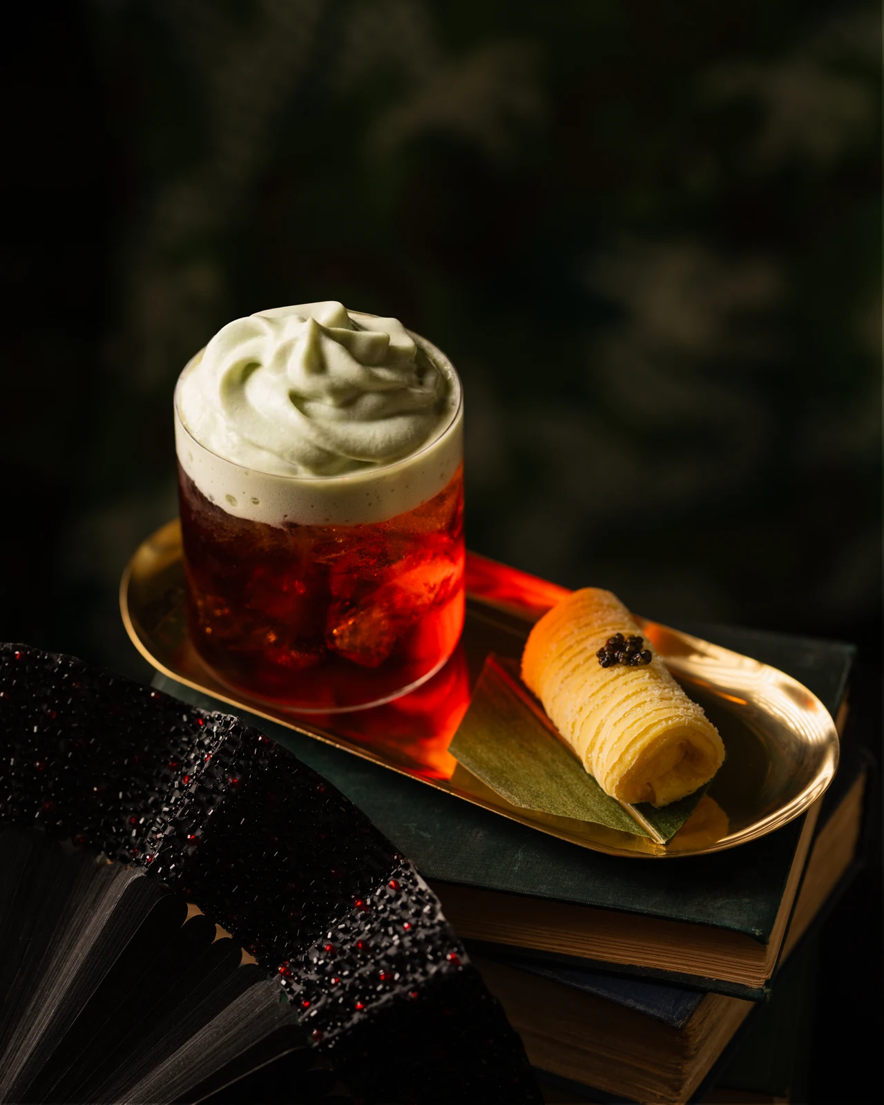 Red cocktail topped with pale green foam beside a pastry and black beaded fan