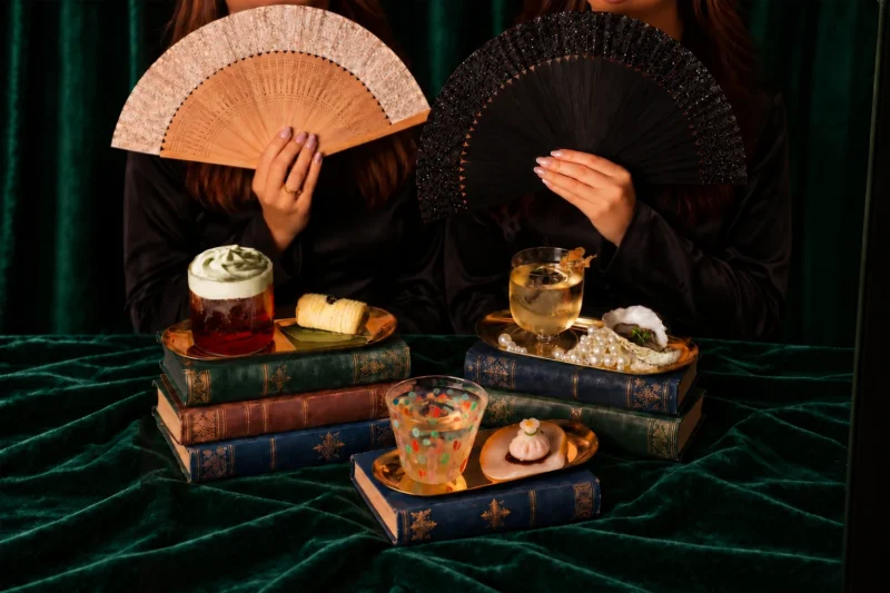 Three cocktail and canapé pairings arranged on books against green velvet, with beaded fans