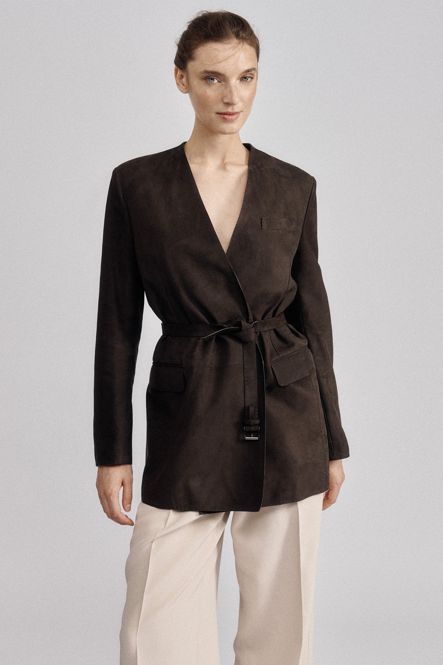 Model wearing a dark brown belted jacket with a deep V neckline and pale trousers.