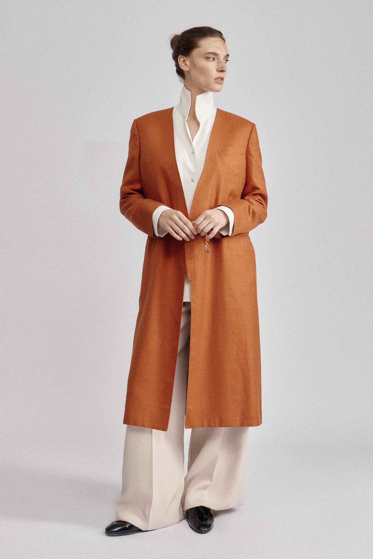 Full-length model wearing an orange long coat over a white blouse and wide pale trousers.