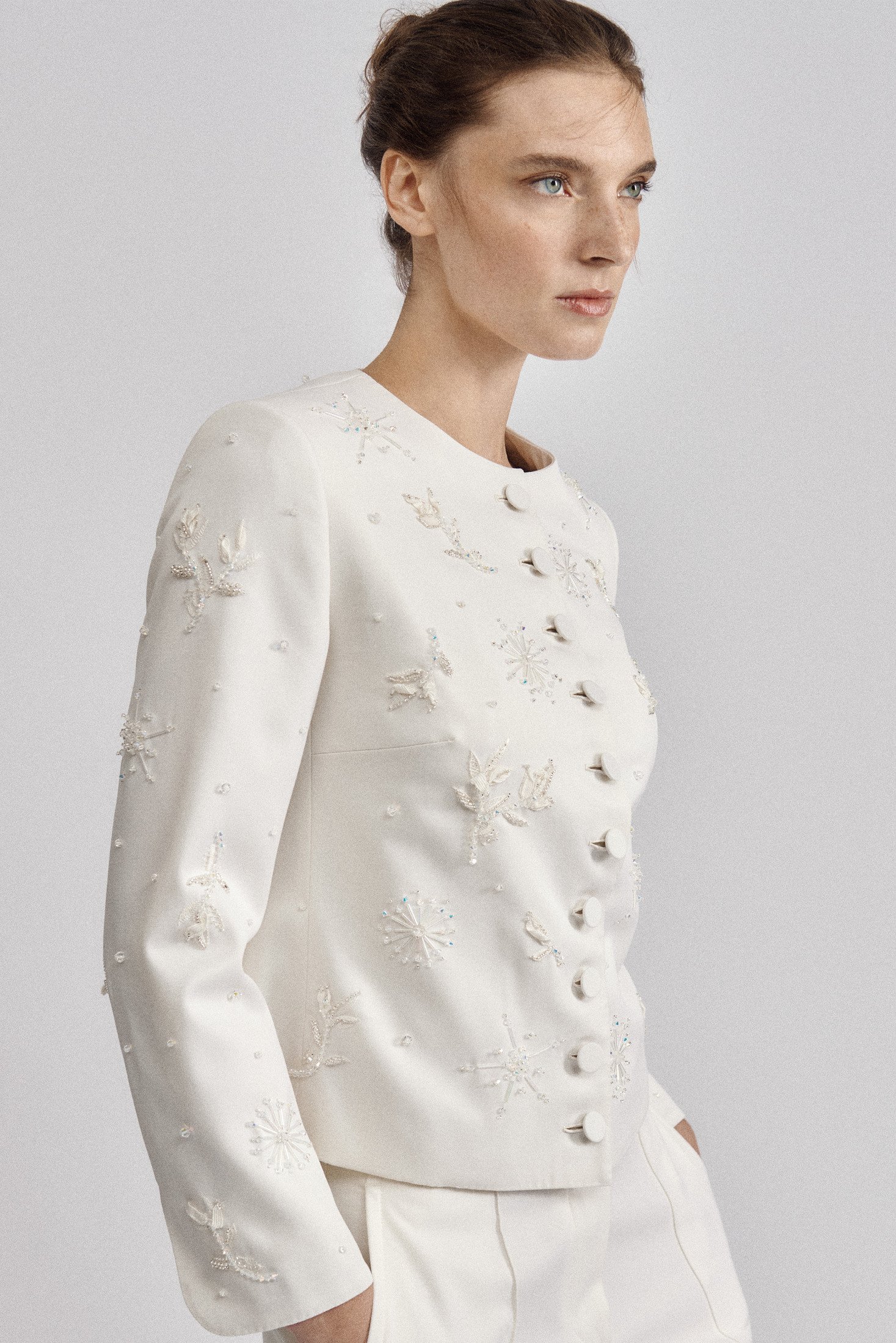 Close view of a model wearing a white collarless jacket with raised leaf-like embroidery and white trousers.