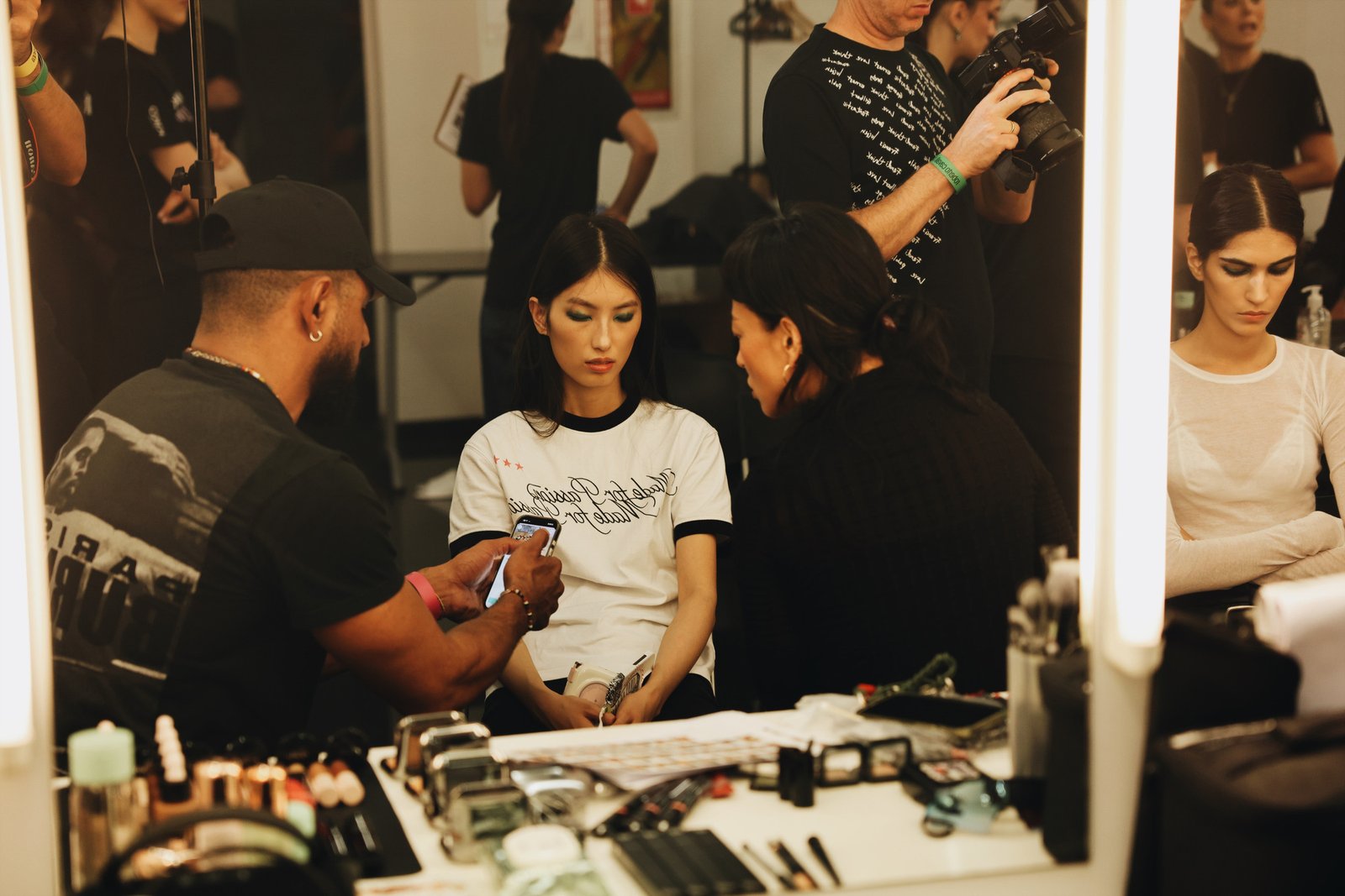 Models and makeup artists at a brightly lit backstage dressing table.