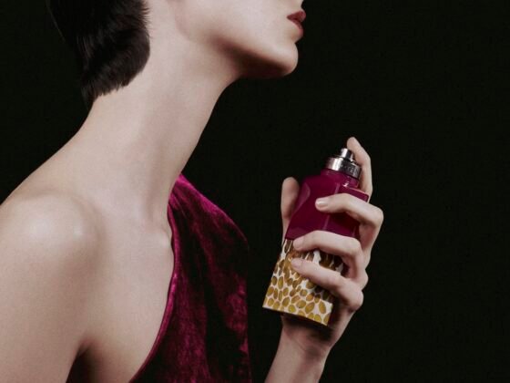 A cropped profile of a model holding a purple perfume bottle with a golden seed-patterned base against a dark background