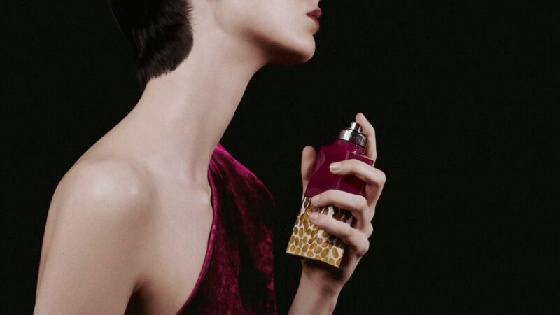 A cropped profile of a model holding a purple perfume bottle with a golden seed-patterned base against a dark background