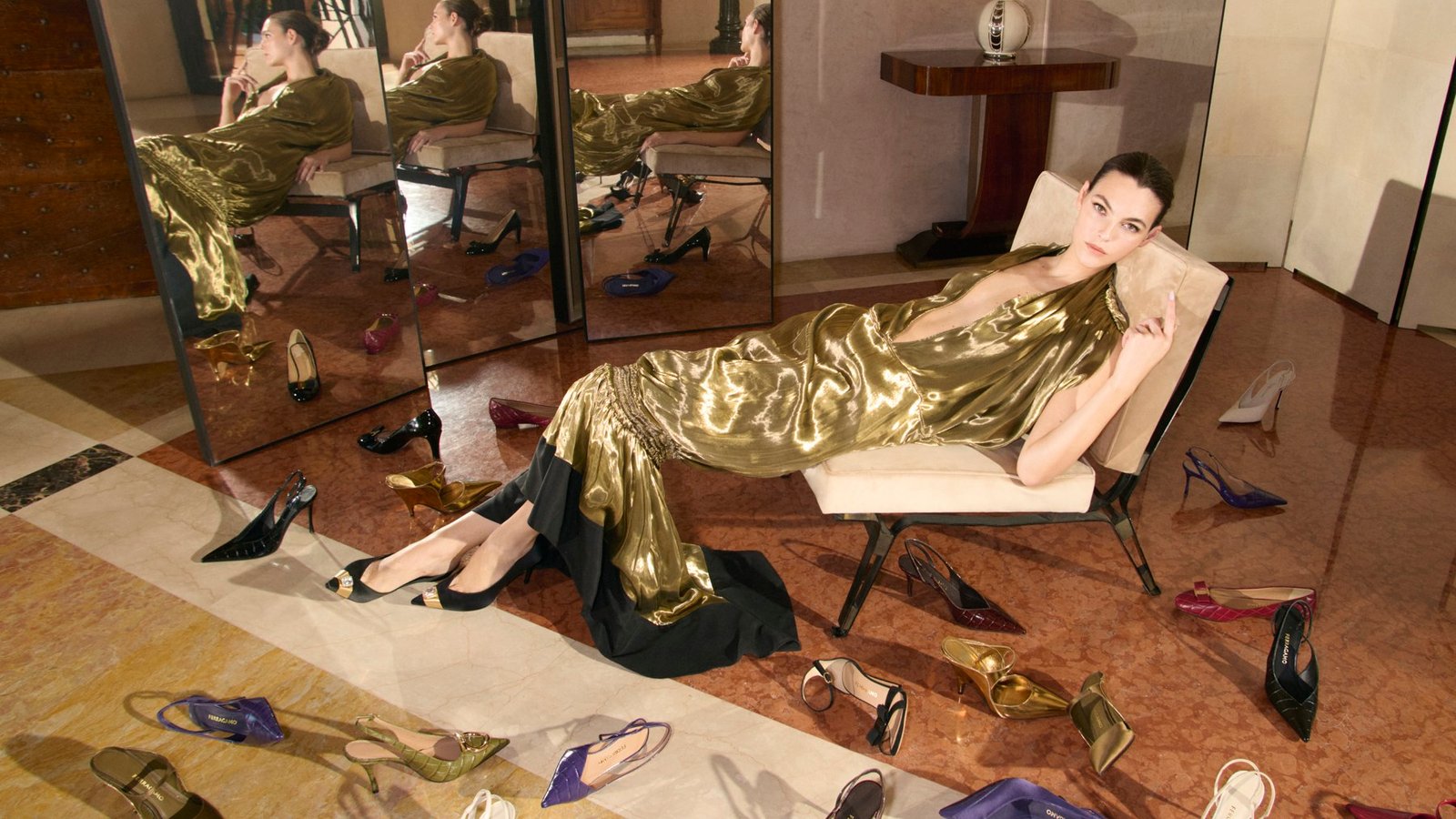A model in a draped gold-coloured dress reclines before folding mirrors, surrounded by scattered Ferragamo shoes
