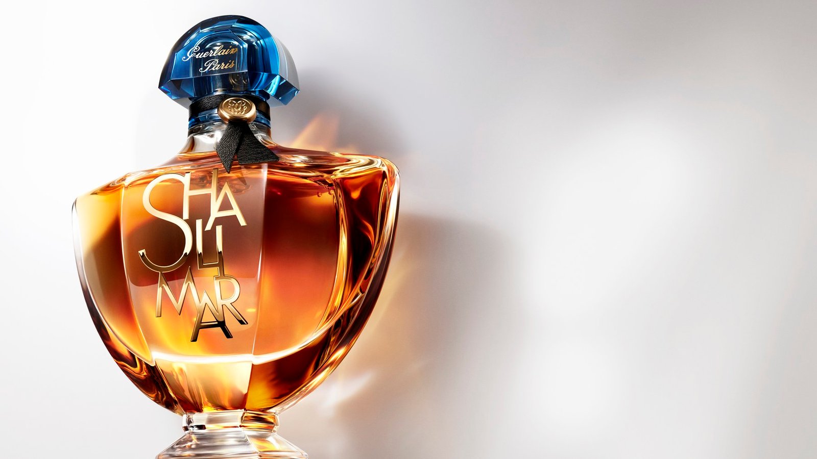 An amber Shalimar L’Essence bottle with a blue fan-shaped cap and stacked golden lettering, lit from the side