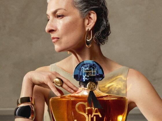 A grey-haired woman in profile poses with an oversized Shalimar L’Essence bottle against a warm neutral background
