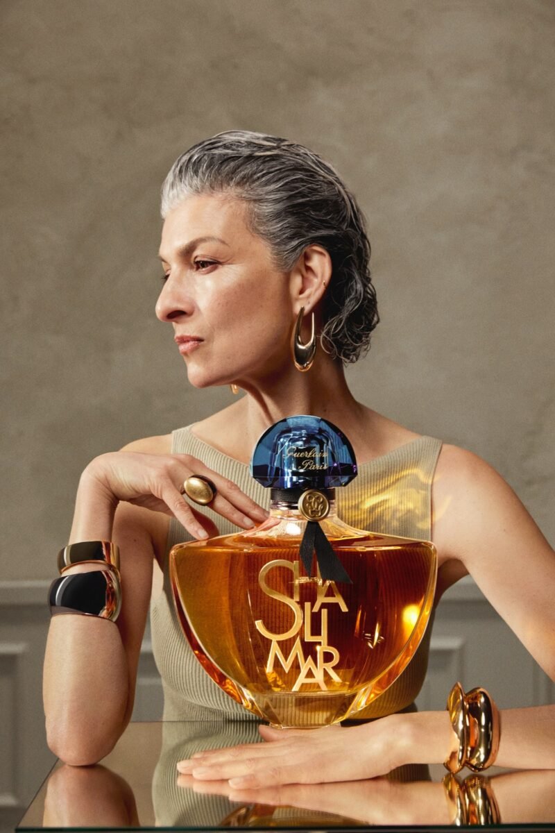A grey-haired woman in profile poses with an oversized Shalimar L’Essence bottle against a warm neutral background