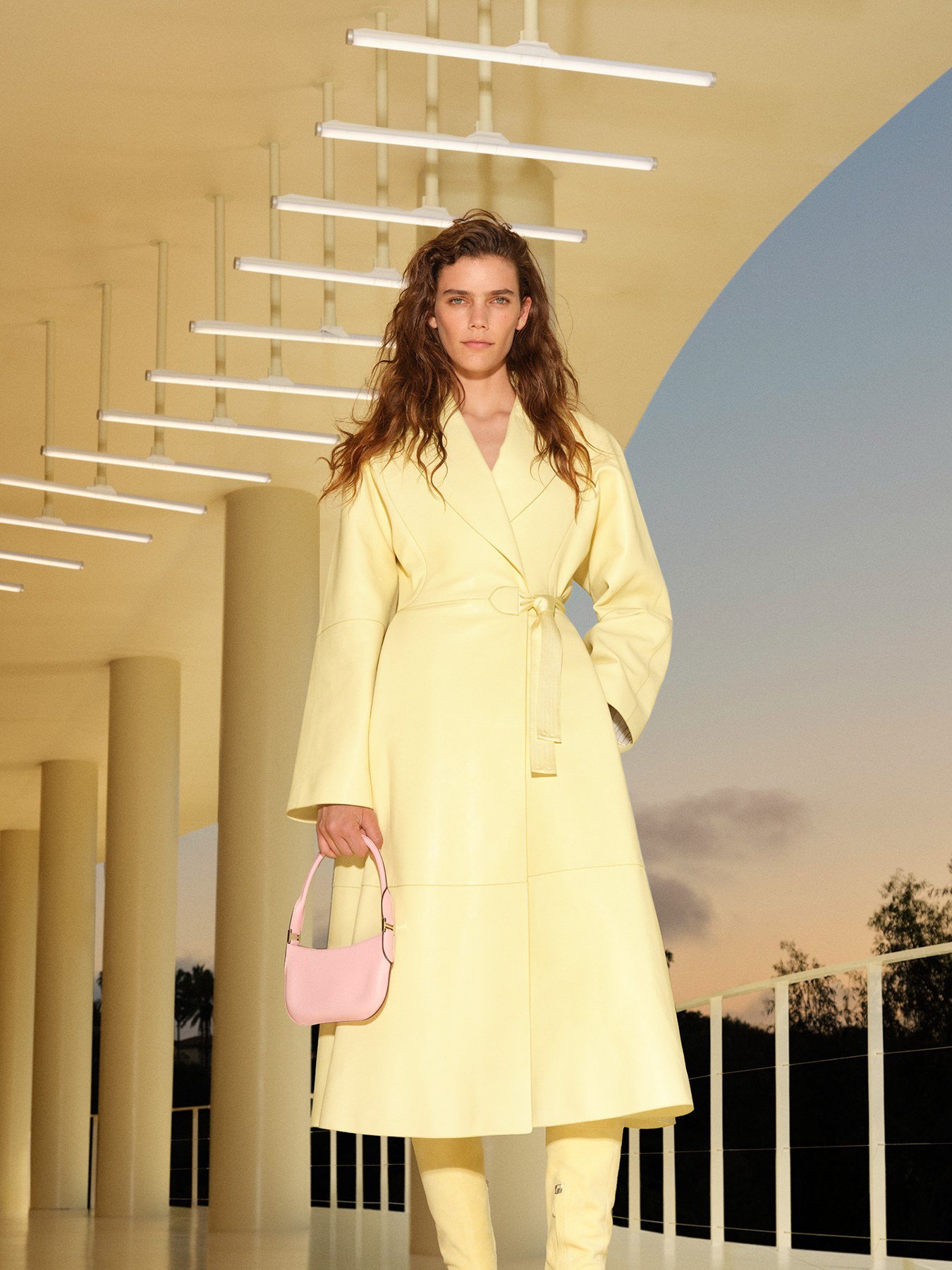 Model in a pale yellow Hermès coat carrying a pink shoulder bag beneath a lit pavilion