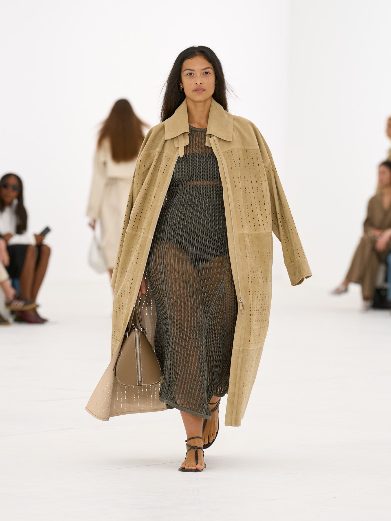 Hermès SS27 runway look with perforated tan coat over a dark open-knit dress