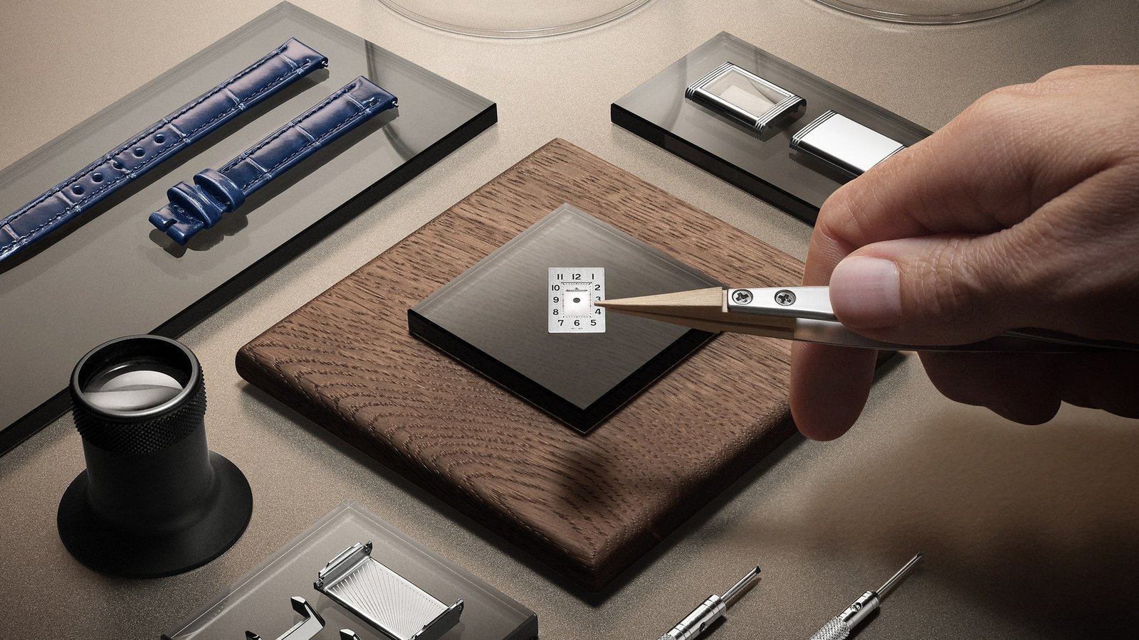Tweezers beside a Reverso Origin dial, with steel case parts and a blue strap arranged nearby