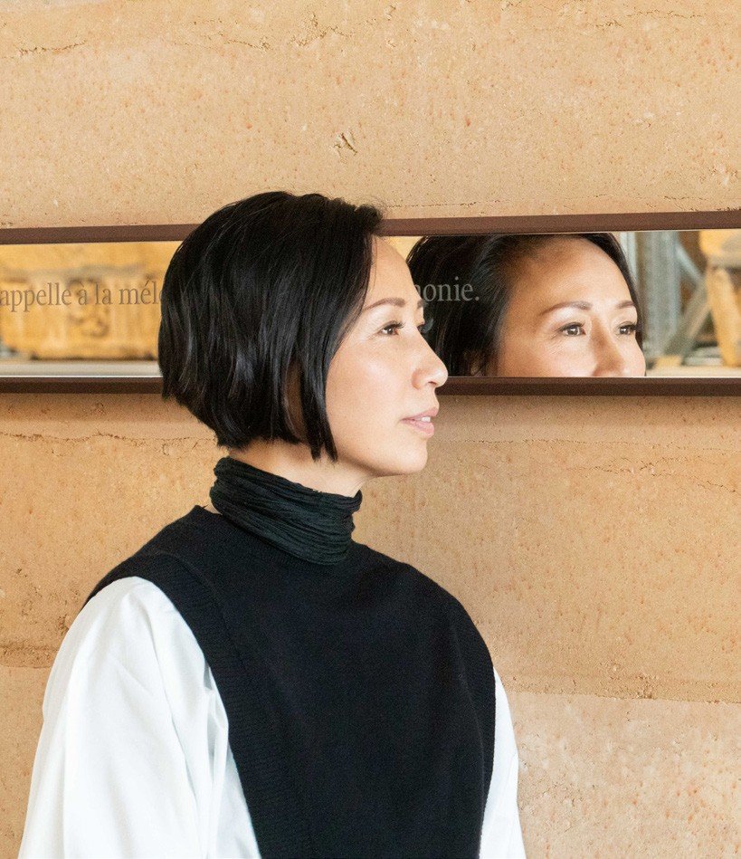 Jiang Qiong Er in profile wearing a black sleeveless top over a white shirt, beside a narrow wall mirror