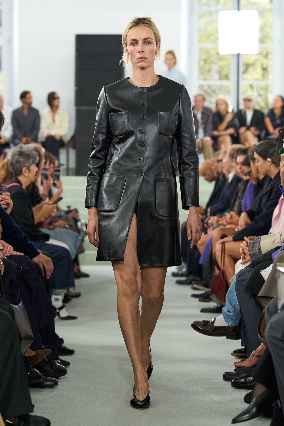 A model in a glossy black collarless coat with chest pockets, a buttoned front and an open lower panel, worn with pointed black shoes