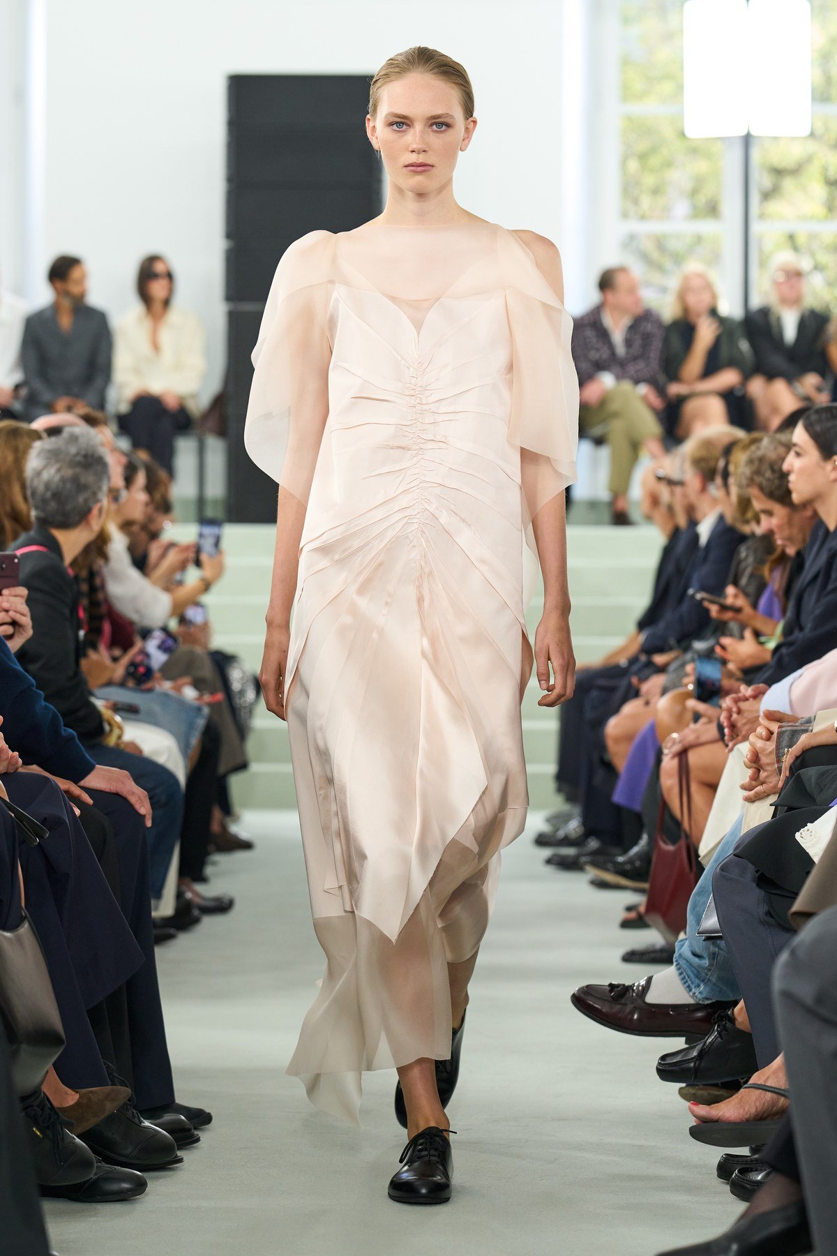 A model wears a pale blush dress with gathered folds through the body and an irregular, flowing hem