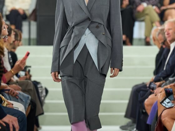 A model wears grey tailoring with angular overlapping front panels and knee-length lower layers over purple tights