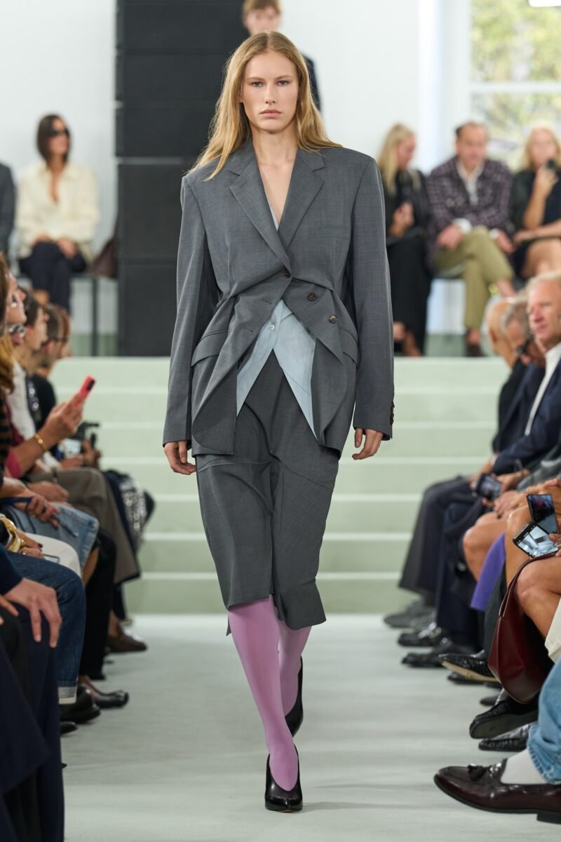 A model wears grey tailoring with angular overlapping front panels and knee-length lower layers over purple tights