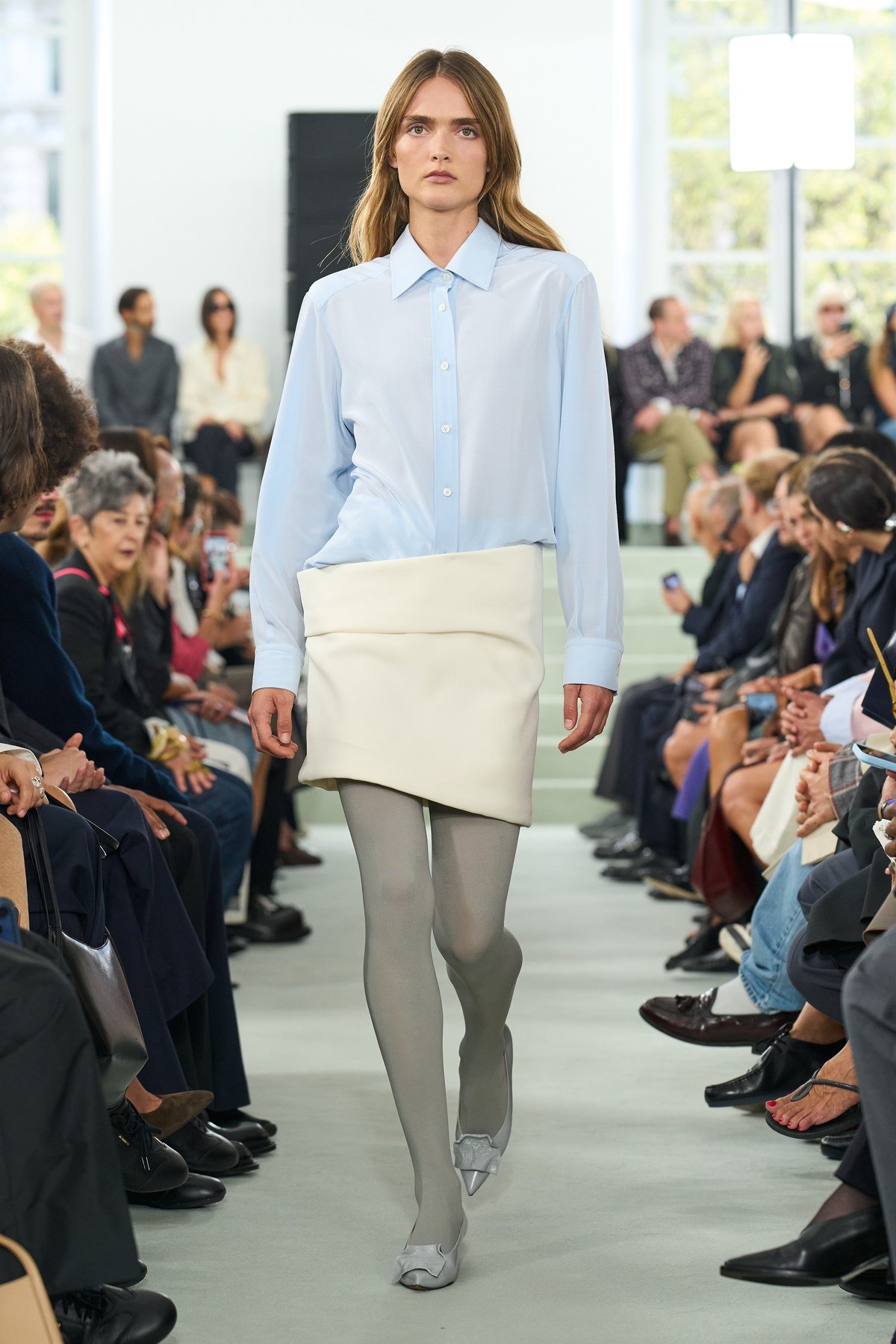 A model wears a pale blue shirt above a short cream skirt with a broad folded upper edge, grey tights and pointed shoes