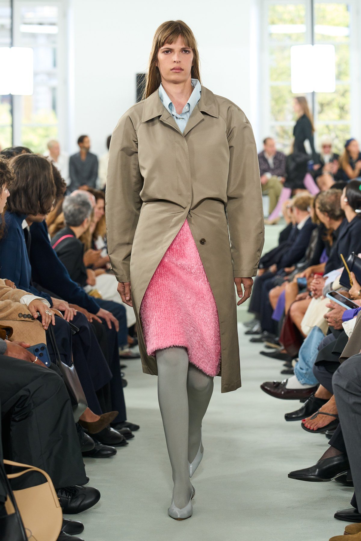 A model wears a khaki-toned coat gathered at the waist, opening over a bright pink textured panel and grey legwear