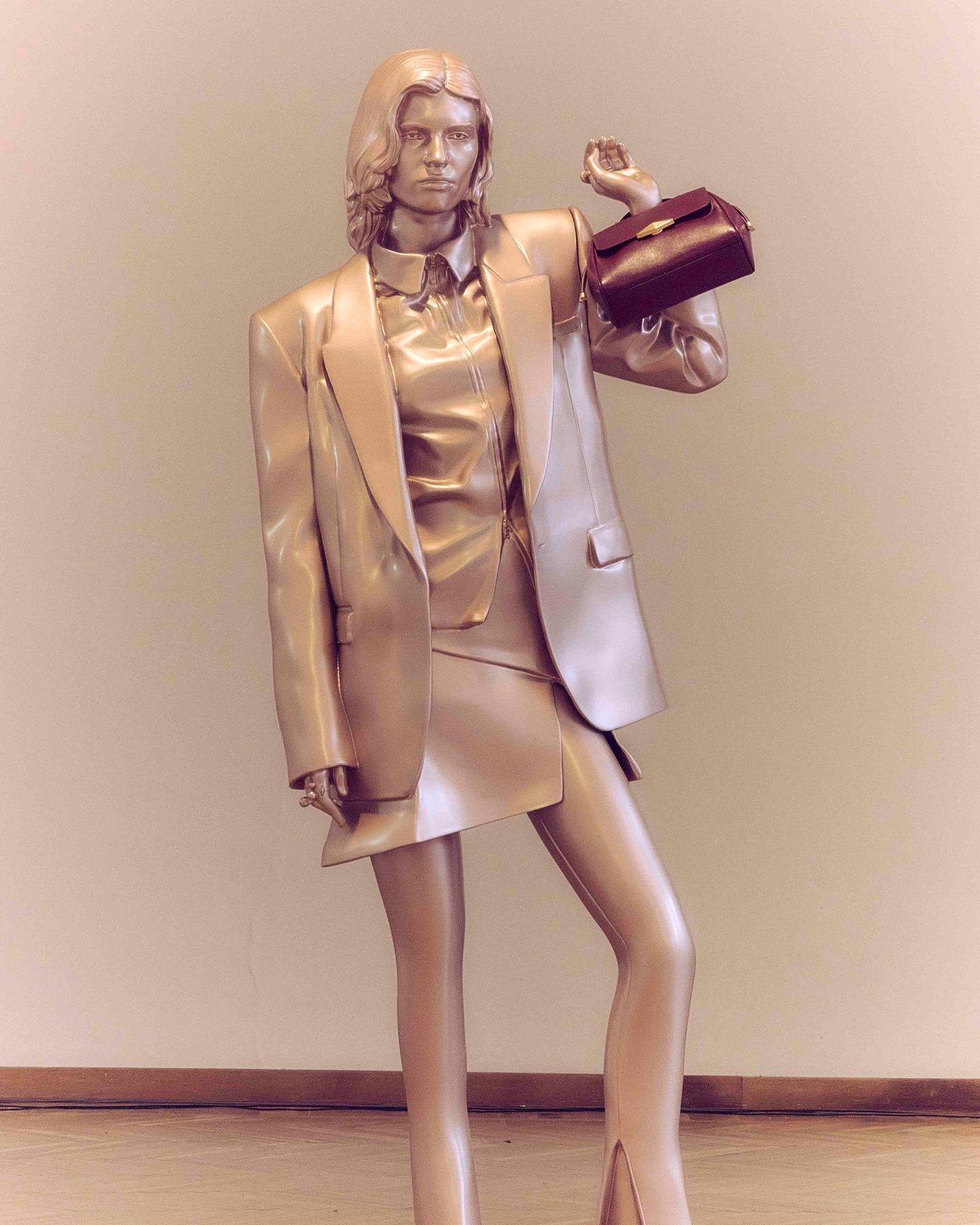 A life-size gold-toned figure in a tailored jacket holds a deep burgundy handbag aloft in the collection presentation