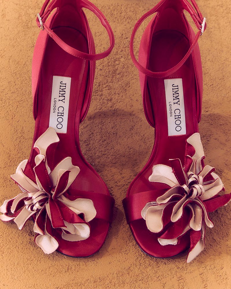 A pair of ruby-red open-toe heels with ankle straps and large layered red-and-pale-pink floral corsages, viewed from above