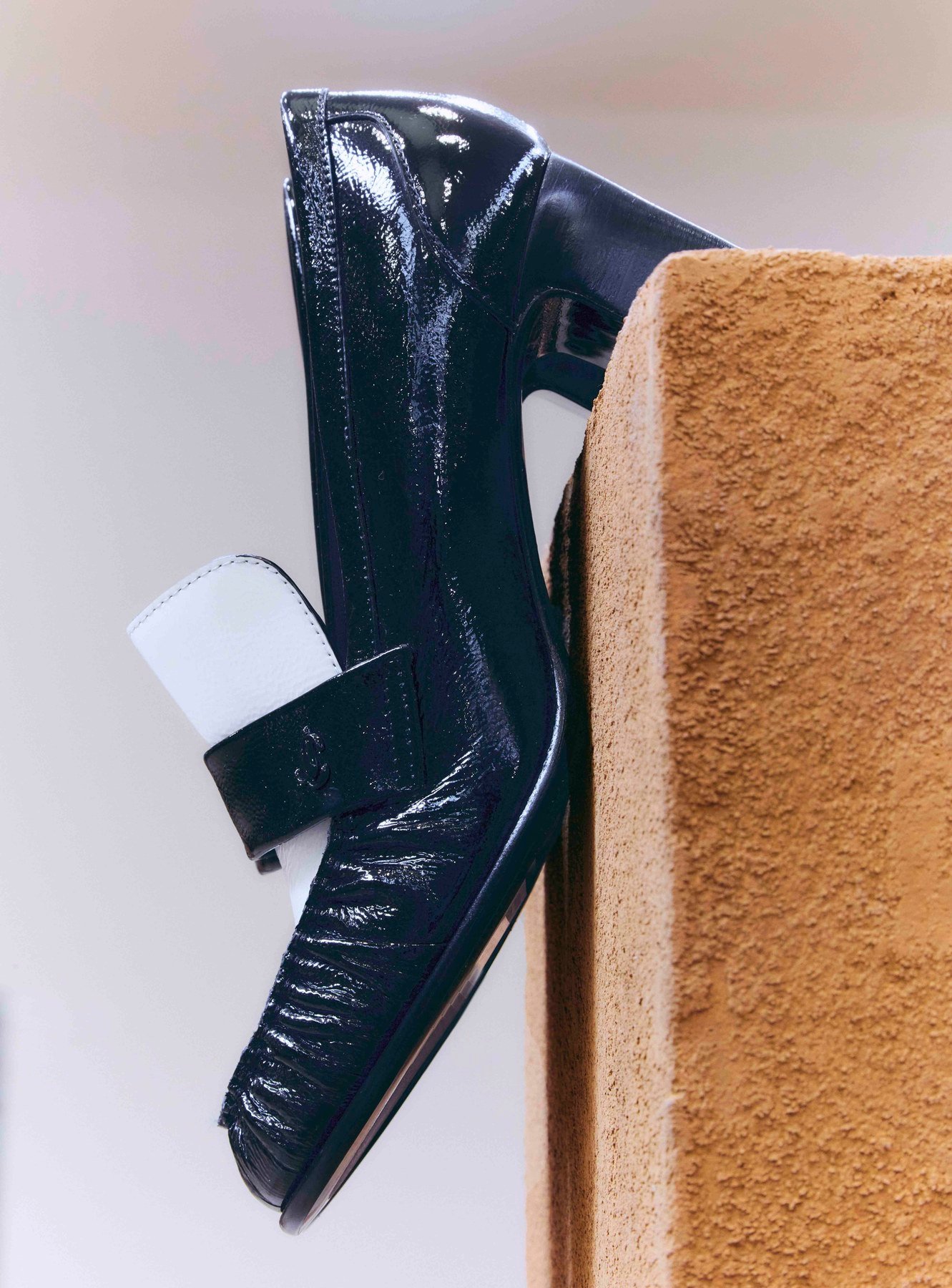 Close side view of a glossy black loafer with a contrasting cream panel and angular block heel, resting beside a textured ochre plinth