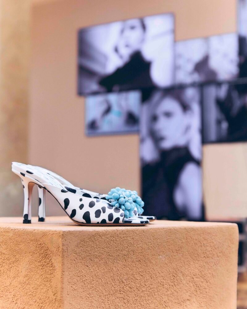 A black-and-white spotted peep-toe mule with a turquoise-coloured bead cluster, displayed on an ochre plinth before black-and-white portraits