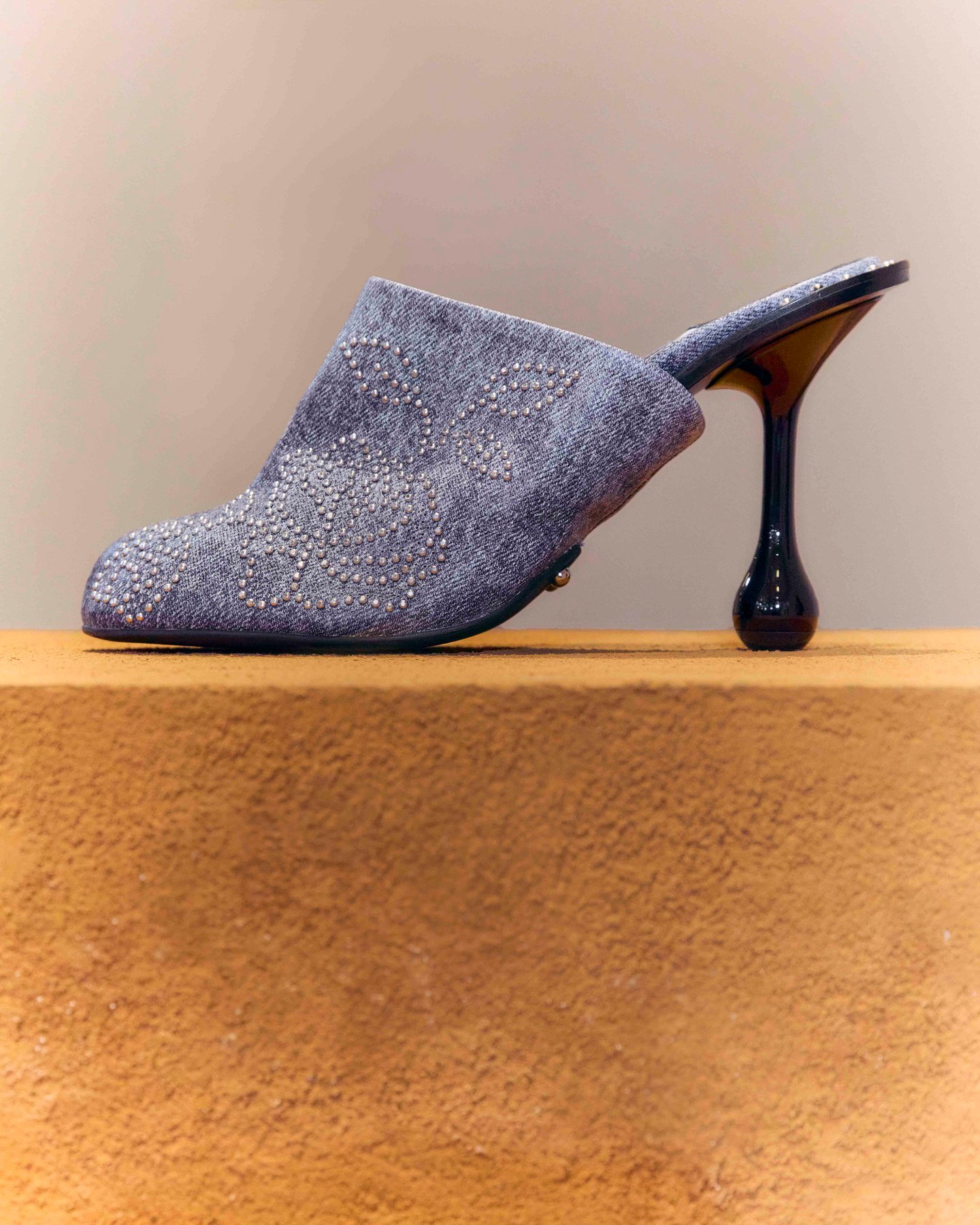 A blue denim-effect mule with a fine studded floral motif and a curved black heel, displayed on an ochre plinth