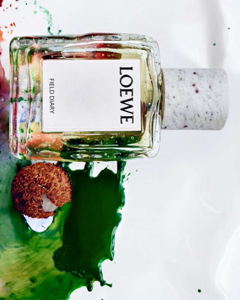 Loewe Field Diary bottle beside lychee and green pigment in a textured still life