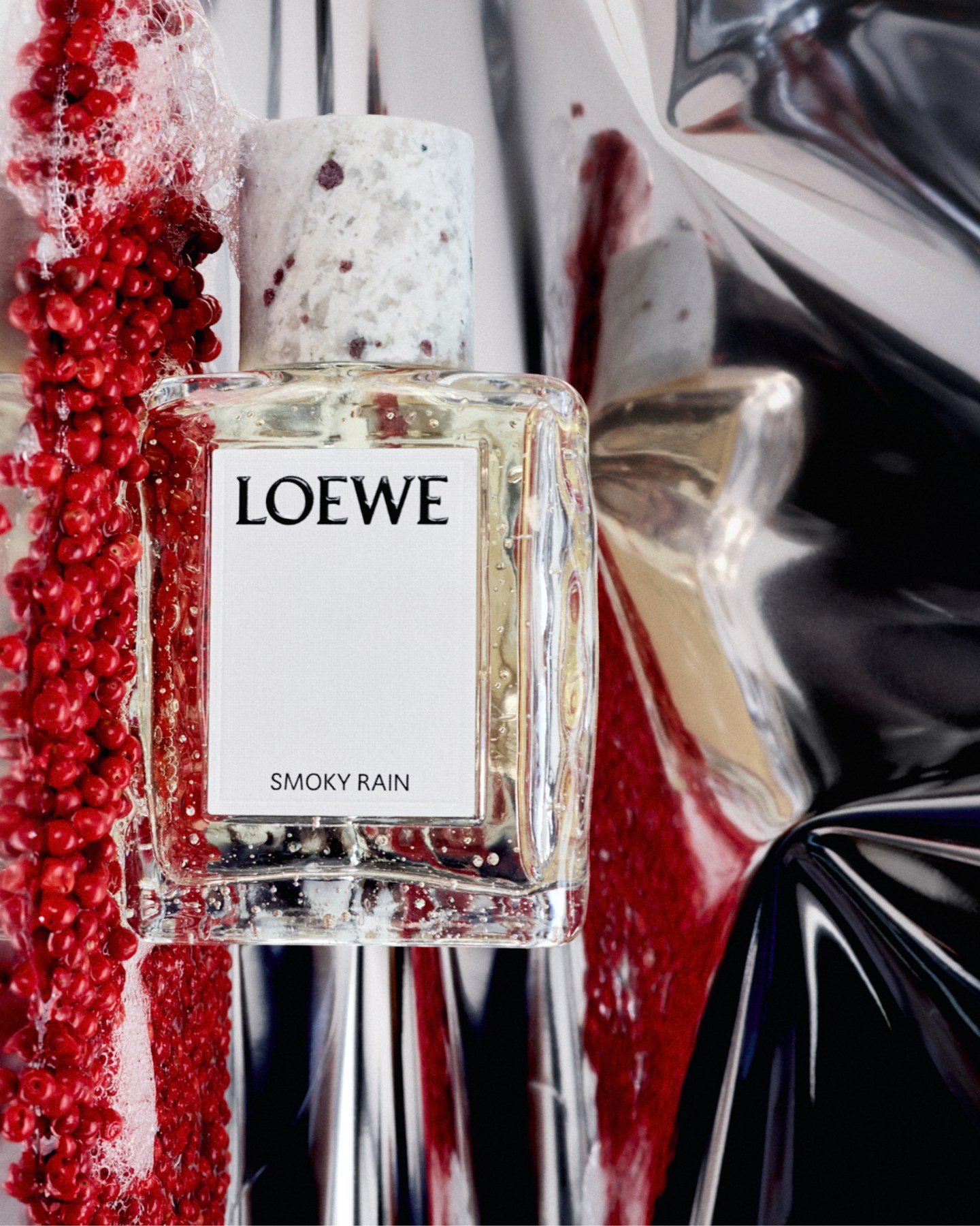 Loewe Smoky Rain bottle framed by glossy red peppercorns and reflective silver surfaces