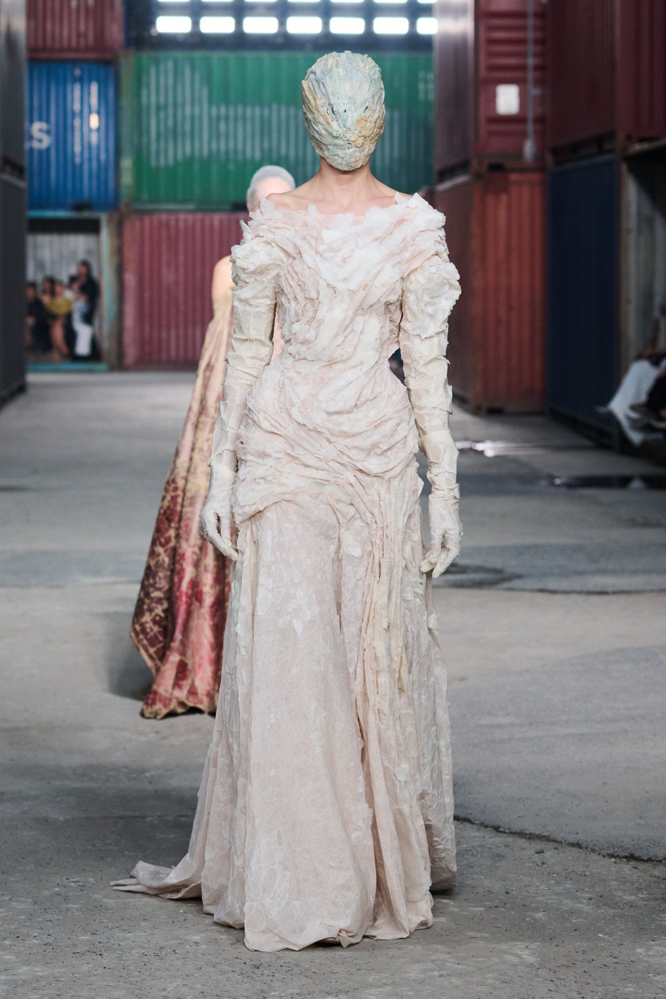 Front view of a pale full-length dress with rough layered folds around the shoulders, torso and sleeves, worn with an Artisanal mask.