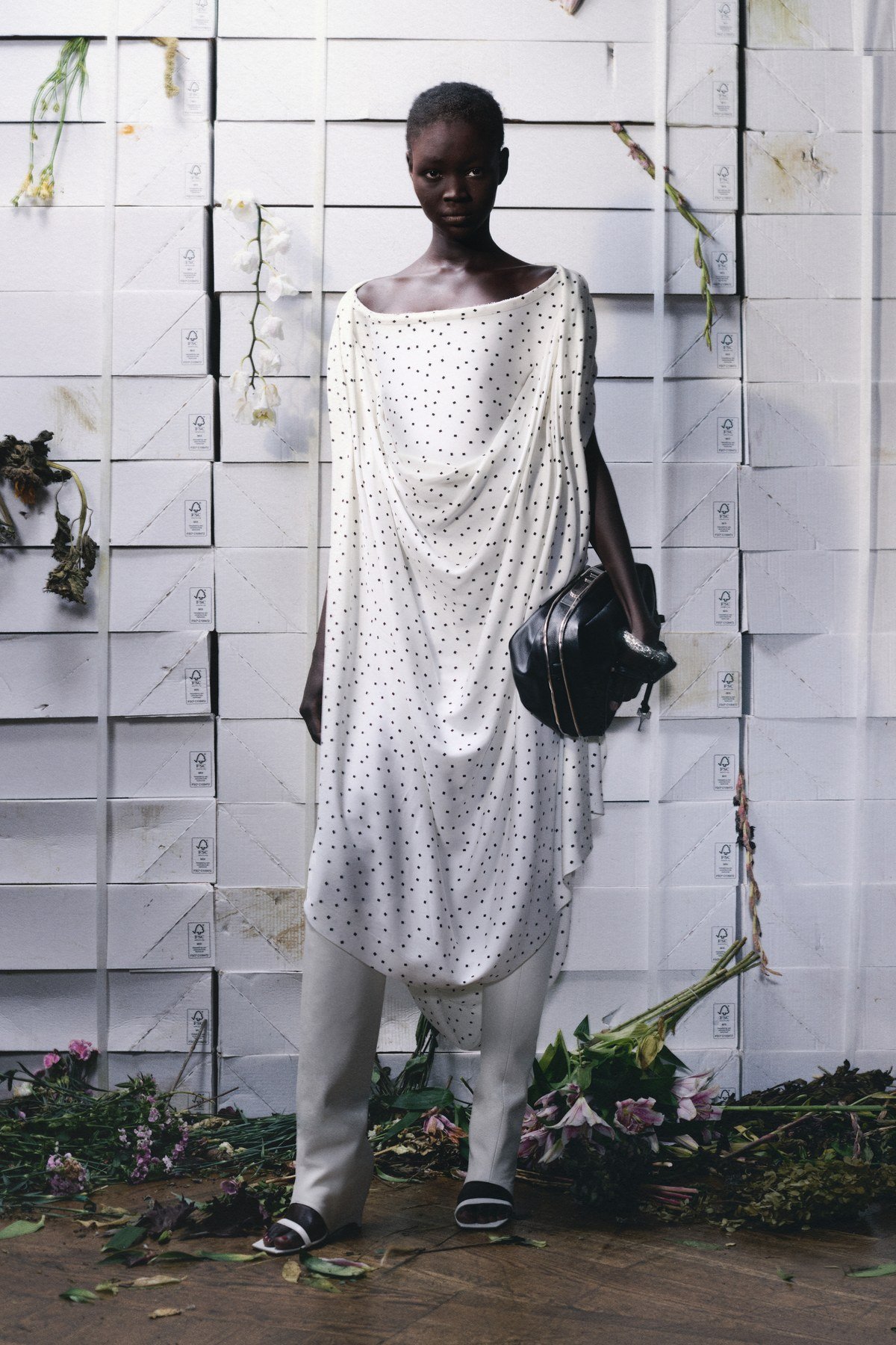 A model wears a white dress scattered with small black squares, draped from a curved neckline above white open-toe boots