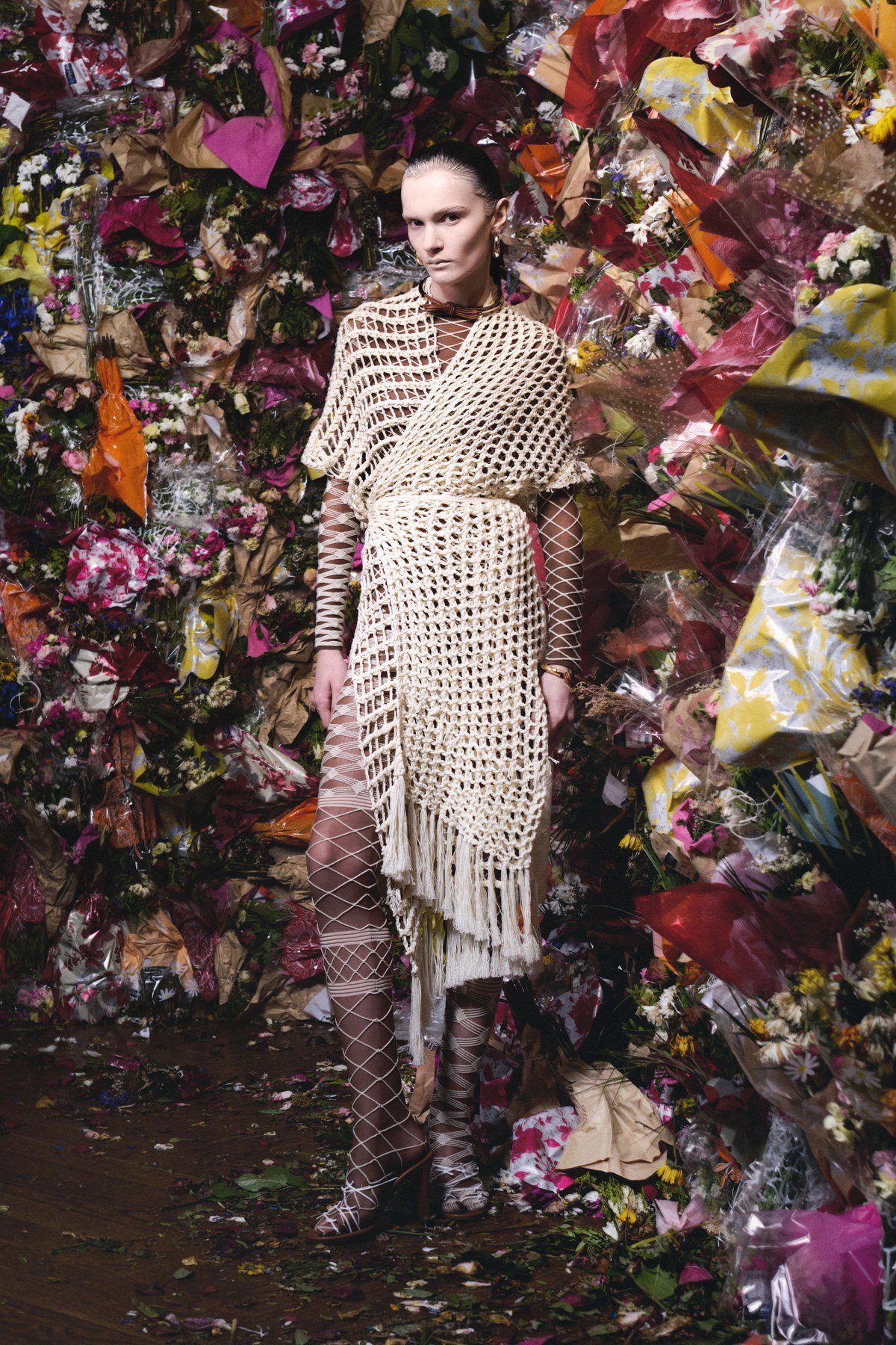 A model in an open cream crochet layer tied at the waist, over net-patterned second-skin pieces, against a dense floral backdrop