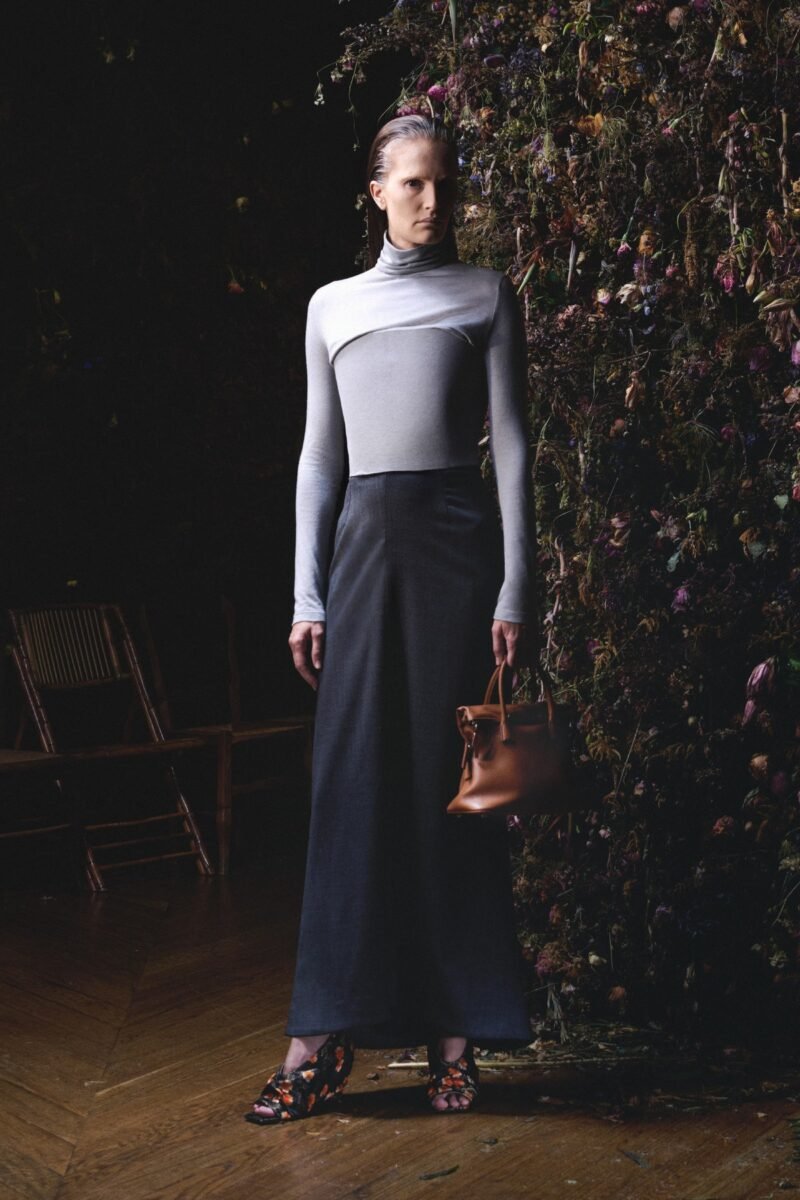 A model wears a pale grey high-neck top with a raised curved chest panel and a long dark skirt, beside an aged floral wall