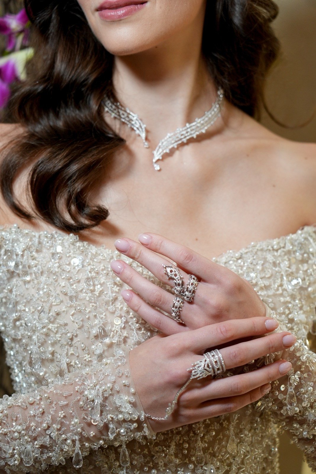 Close-up of a branching necklace, rings and a hand chain against an off-the-shoulder gown covered in pale embellishment.