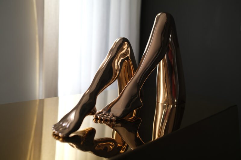 Two polished bronze sculptures of bent legs reflected in a dark tabletop