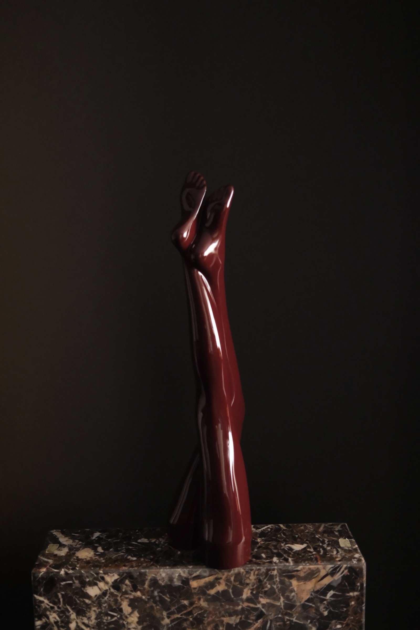 Dark red bronze leg sculpture standing on a brown-veined stone plinth against a dark background