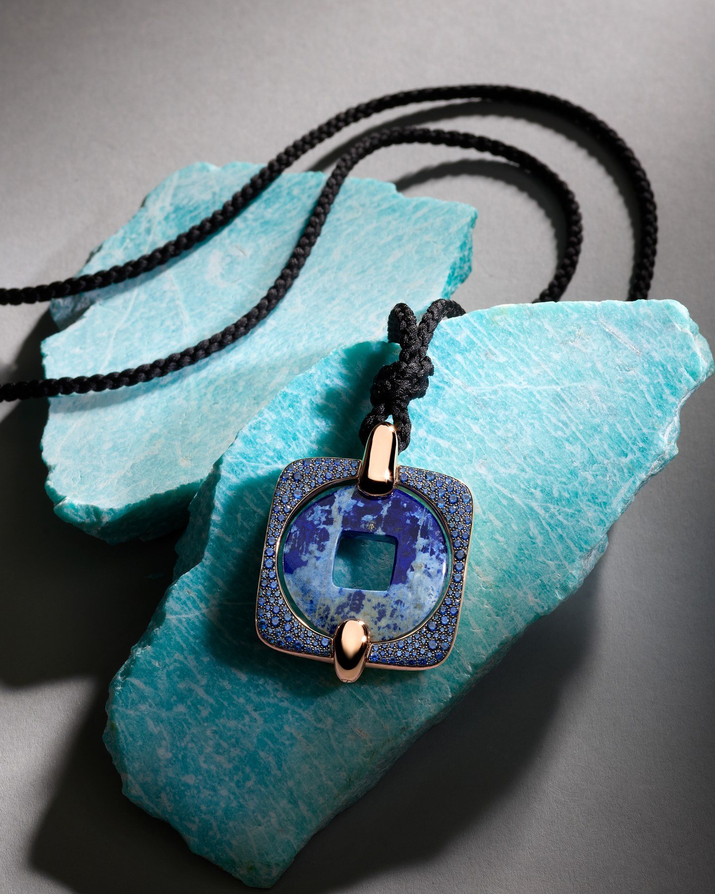 Blue Moneta Orientale pendant on a black cord against a turquoise-coloured mineral specimen