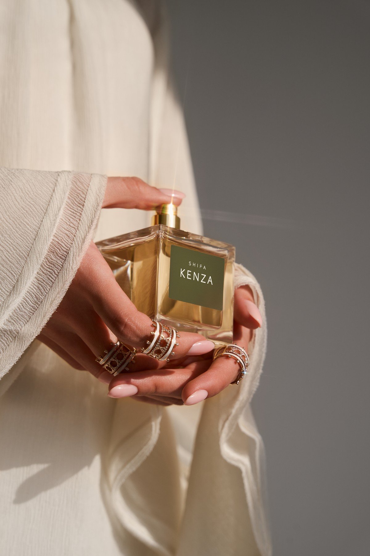 Hands wearing openwork rings hold a Shifa Kenza perfume bottle against ivory sleeves
