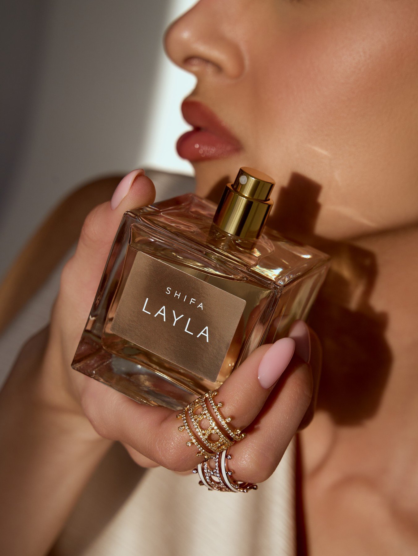A model holds a square Shifa Layla perfume bottle beside her face, with rings visible on her fingers
