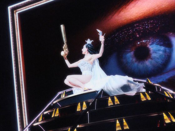 Dita Von Teese in white costume on a theatrical platform beneath a projected eye