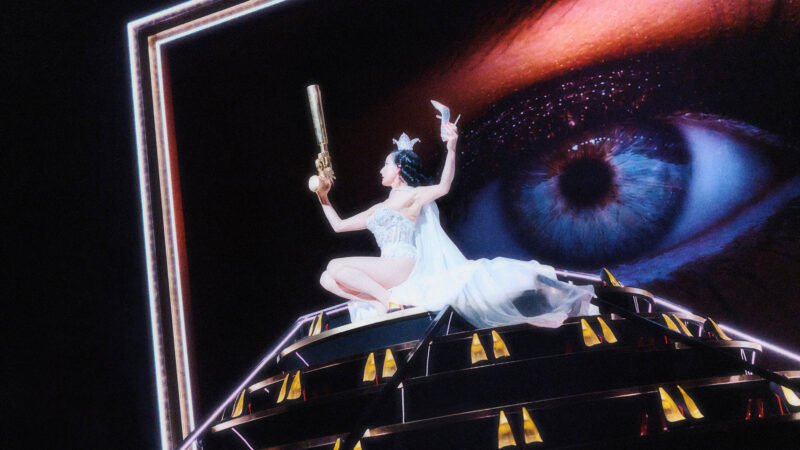 Dita Von Teese in white costume on a theatrical platform beneath a projected eye