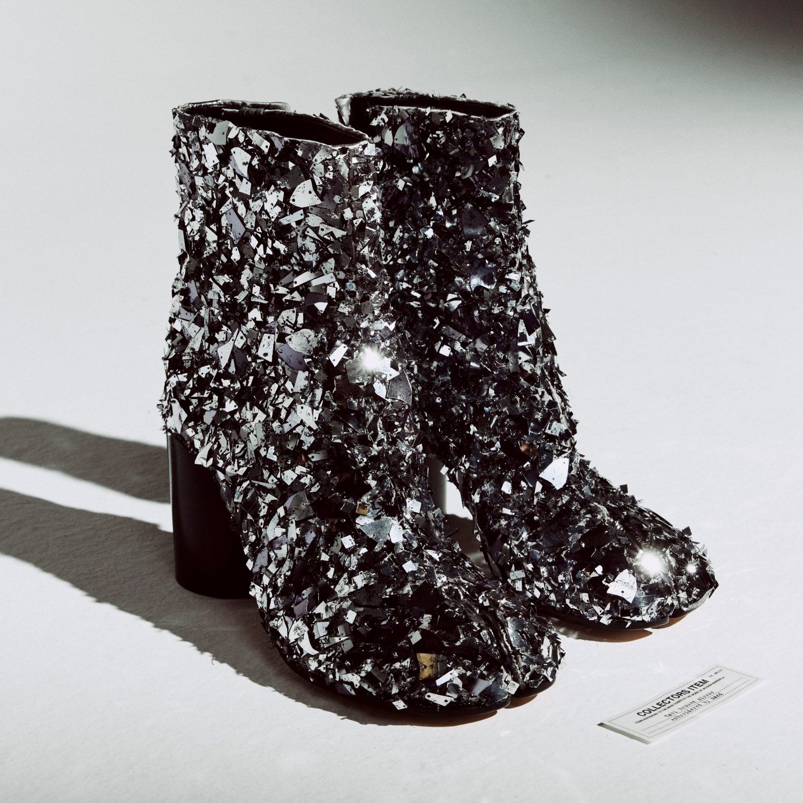 A pair of split-toe ankle boots densely covered in small reflective silver-toned embellishments, with dark cylindrical heels