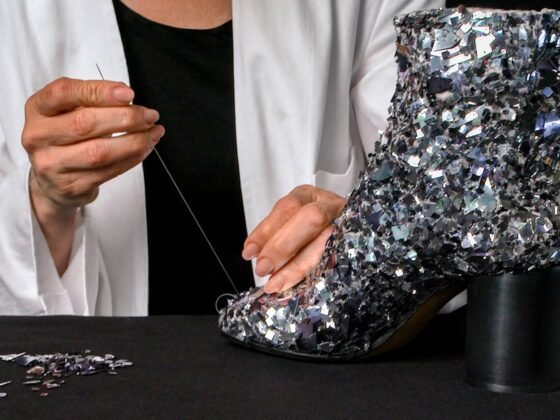 A craftsperson in a white coat draws thread through a silver-toned embellished split-toe ankle boot on a black worktable