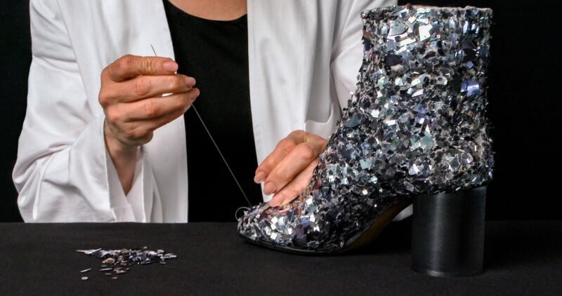 A craftsperson in a white coat draws thread through a silver-toned embellished split-toe ankle boot on a black worktable