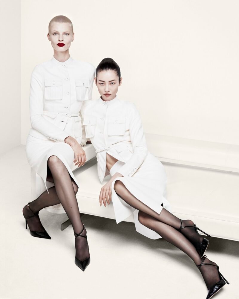 Two models in white belted shirt dresses and sheer black hosiery pose on a white bench, wearing pointed Tom Ford heels