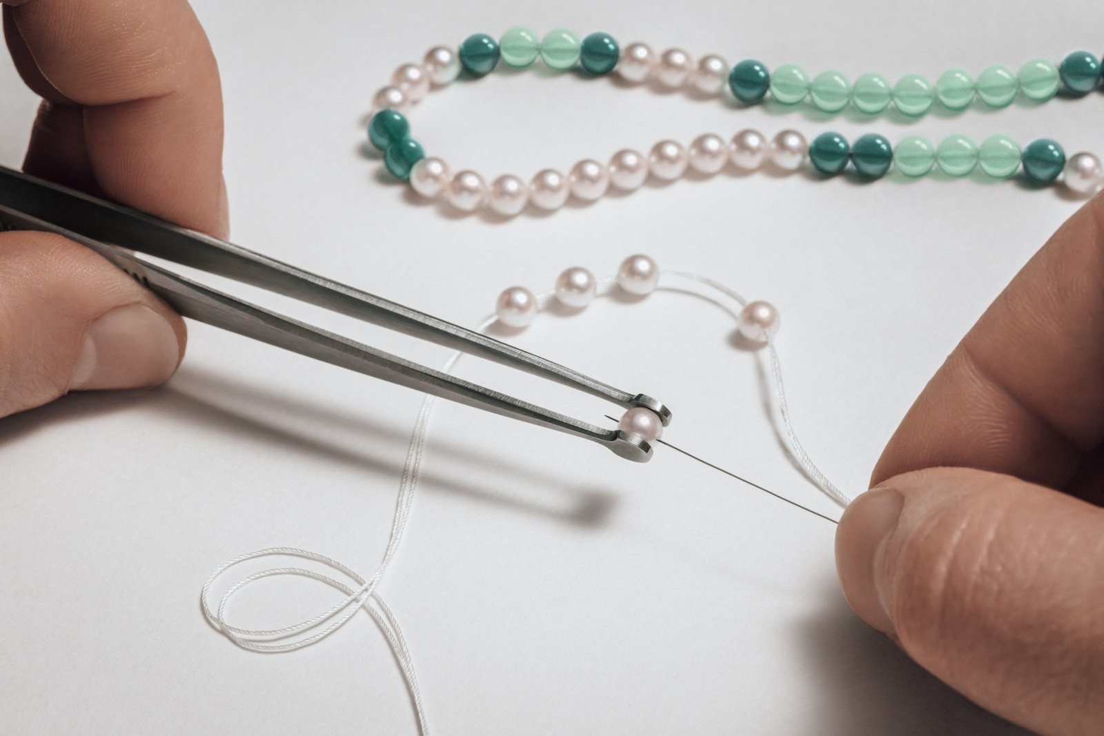 Hands and tweezers threading a pearl beside green stone beads and Akoya pearl strands