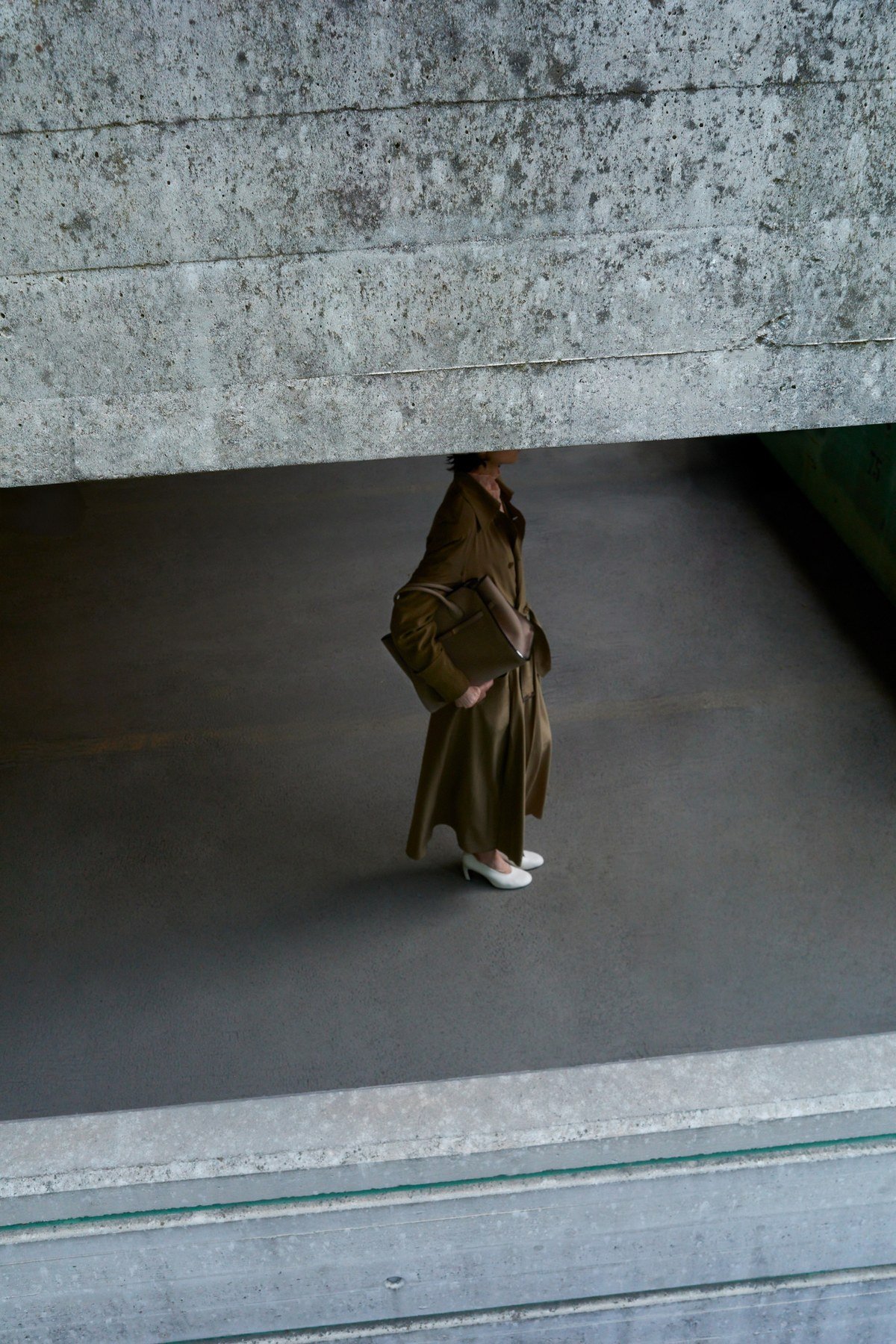 A model carrying a brown bag walks beneath a broad concrete structure in a high-angle architectural photograph