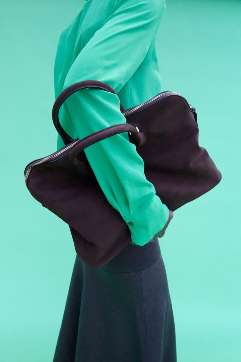 A soft dark bowling bag loops over the arm of a model wearing a vivid green top against a turquoise background